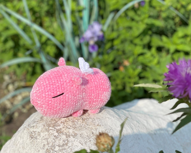 K&ouml;nnte beinhalten: Ein rosa geh&auml;keltes Stofftier mit wei&szlig;en Fl&uuml;geln. Das Tier liegt auf einem wei&szlig;en Stein mit gr&uuml;nen Pflanzen im Hintergrund.