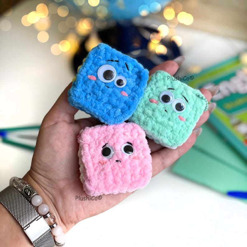 May include: Three handmade, plush, square-shaped toys in blue, green, and pink. Each has googly eyes and embroidered details, with a smiling or sad expression. The toys are held in a hand, with a bracelet visible in the foreground.
