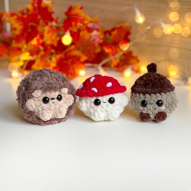 May include: Three crocheted plush toys: a brown hedgehog, a mushroom with a red cap and white spots, and a brown acorn. Each toy has black eyes and a smiling face. The background features autumn leaves and soft lighting.