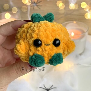 May include: A plush, smiling pumpkin with large black eyes, a green stem, and green feet. The pumpkin is made of soft, textured orange fabric. The product is a handmade item.