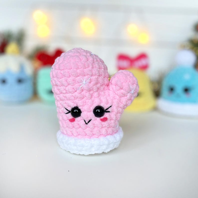 May include: A pink crocheted mitten plush toy with a white cuff, featuring black plastic safety eyes, embroidered eyelashes, a small V-shaped mouth, and two pink stitched cheeks. The mitten has a soft, textured yarn surface.