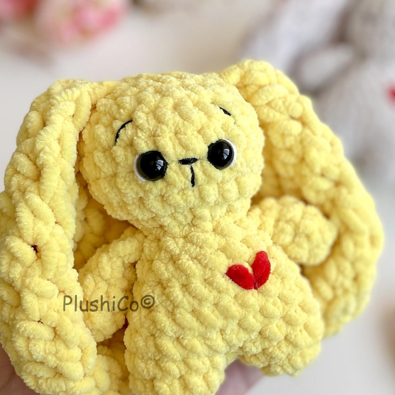 May include: A yellow crocheted bunny with large black eyes and a red heart on its chest. The bunny has long, braided ears.
