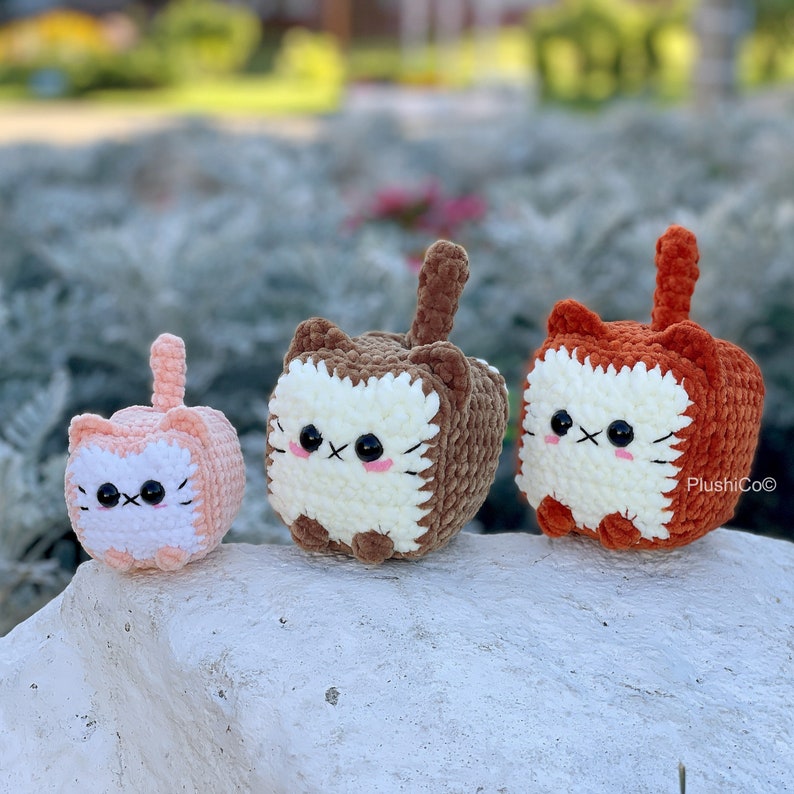 May include: Three crocheted plush cat toys in different colours and sizes. The largest cat is orange, the middle cat is brown, and the smallest cat is pink.