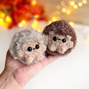 May include: Two handmade plush hedgehogs. One is light beige, the other is brown. Both have black eyes, pink cheeks, and tan faces and paws. The plush toys are soft and fluffy, with a cute, friendly appearance. The background is blurred with warm colors.