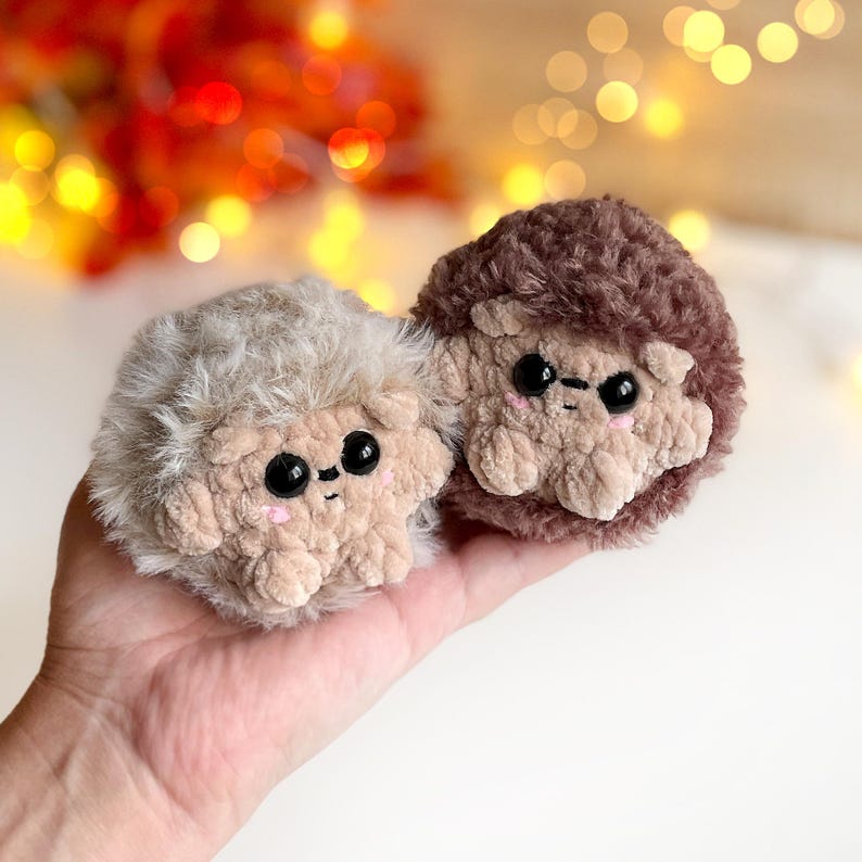 May include: Two handmade plush hedgehogs. One is light beige, the other is brown. Both have black eyes, pink cheeks, and tan faces and paws. The plush toys are soft and fluffy, with a cute, friendly appearance. The background is blurred with warm colors.