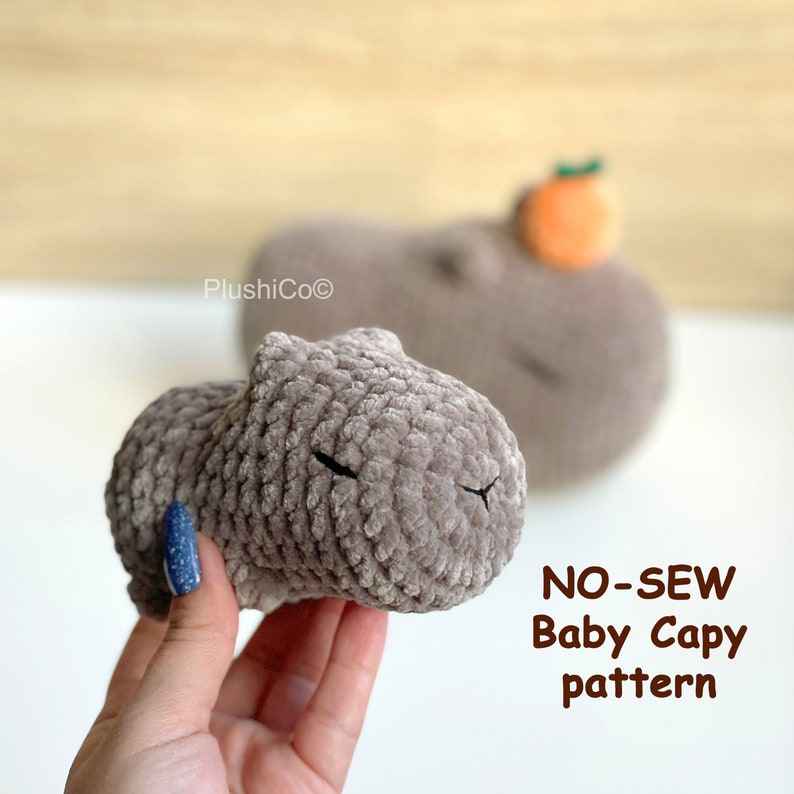 May include: A brown crocheted capybara plush toy with black eyes. The toy is being held in a hand. The text "NO-SEW Baby Capy pattern" is in the lower right corner of the image.
