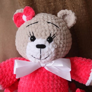 May include: A crocheted teddy bear wearing a pink dress with a white bow. The bear has brown fur, black eyes, and a white nose.