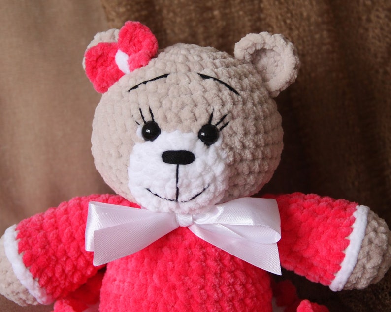 May include: A crocheted teddy bear wearing a pink dress with a white bow. The bear has brown fur, black eyes, and a white nose.