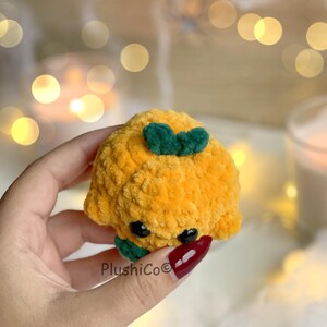 May include: A plush, orange, mushroom-shaped toy with green leaves on top and black eyes. The toy is made of a soft, textured material. The text "PlushiCo" is visible on the bottom left.