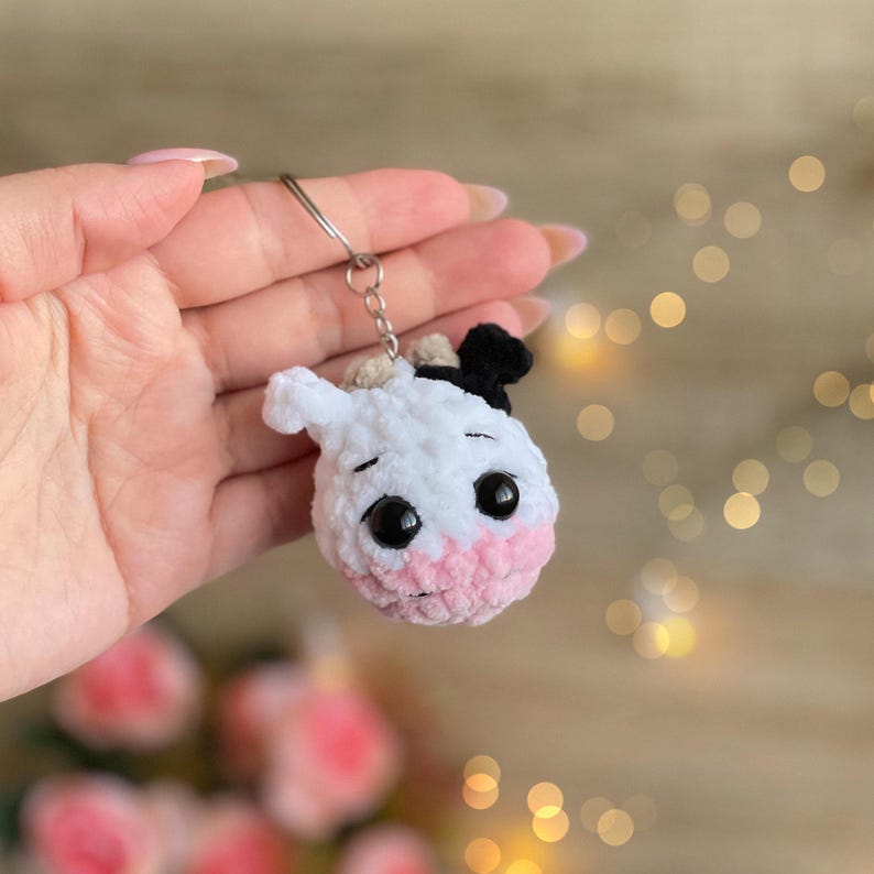 May include: A handmade crochet cow keychain. The cow has a white body, a pink muzzle, and black eyes. It features a small black patch on its head and a silver keyring.