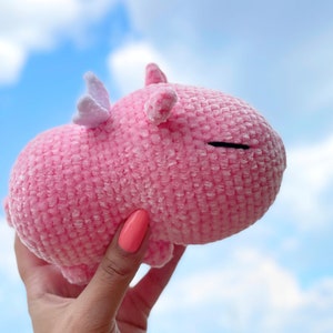 K&ouml;nnte beinhalten: Ein rosa geh&auml;keltes Stofftier mit einem wei&szlig;en Horn und einem geschlossenen Auge. Das Tier wird in einer Hand gehalten.