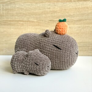 May include: Two gray crocheted capybara plushies. The larger capybara has a small orange crocheted orange on top of its head.