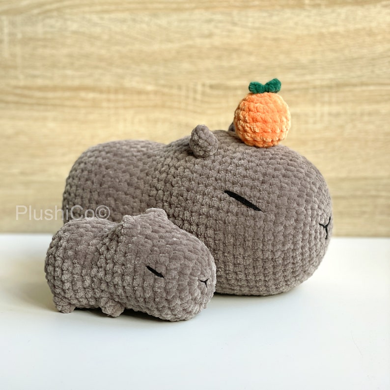 May include: Two gray crocheted capybara plushies. The larger capybara has a small orange crocheted orange on top of its head.