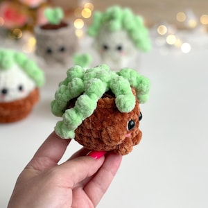 May include: A small, crocheted plush toy shaped like a potted succulent. The base is a textured brown yarn pot, while the top features soft, light green yarn loops resembling plant leaves. It has two black plastic eyes and small pink cheeks.