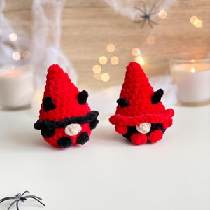 May include: Two small, handmade crochet gnomes featuring bright red pointed hats with black accents. Each gnome has a small beige nose, a black brim, and black details resembling ladybug spots. The figures are crafted from soft yarn.
