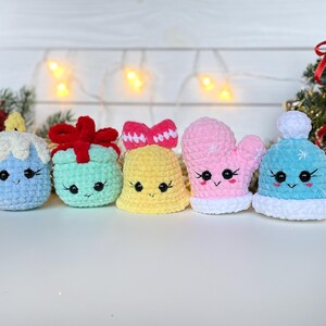May include: Five small crochet plushies featuring cute faces with black eyes and stitched smiles. The set includes a blue candle, a mint green gift with a red bow, a yellow bell, a pink mitten, and a light blue winter hat with a white pom-pom.