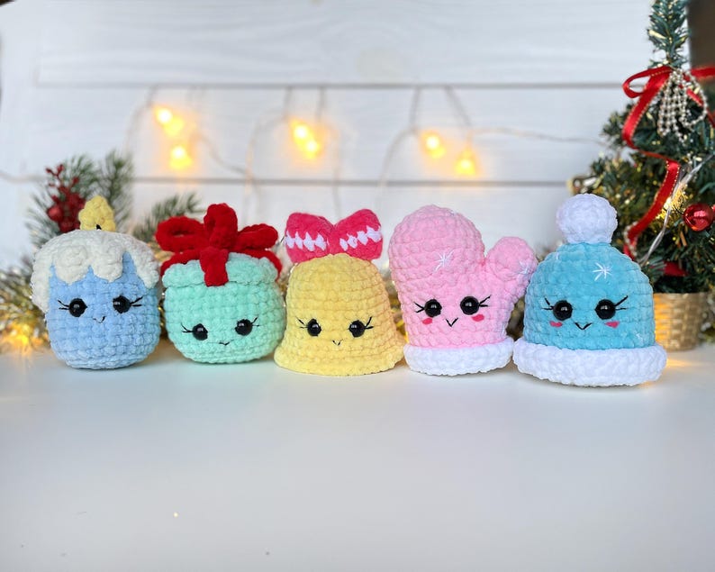 May include: Five small crochet plushies featuring cute faces with black eyes and stitched smiles. The set includes a blue candle, a mint green gift with a red bow, a yellow bell, a pink mitten, and a light blue winter hat with a white pom-pom.