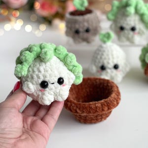 May include: A white and light green crocheted plush toy shaped like a small plant with black bead eyes and pink cheeks, held next to a small, empty, round, brown crocheted pot, with other similar plush plant toys visible in the background.