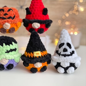 May include: Five plush crochet figures featuring Halloween themes: an orange pumpkin, a red devil, a green Frankenstein-like monster, a black and orange witch, and a white ghost. Each soft, textured character has a round body and a pointed hat.