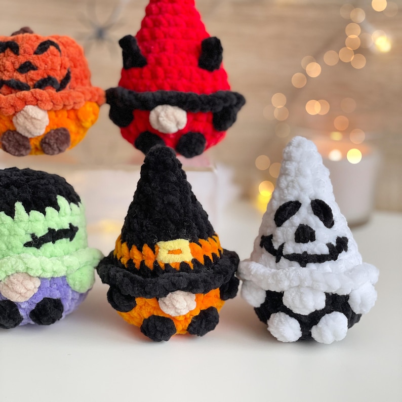 May include: Five plush crochet figures featuring Halloween themes: an orange pumpkin, a red devil, a green Frankenstein-like monster, a black and orange witch, and a white ghost. Each soft, textured character has a round body and a pointed hat.