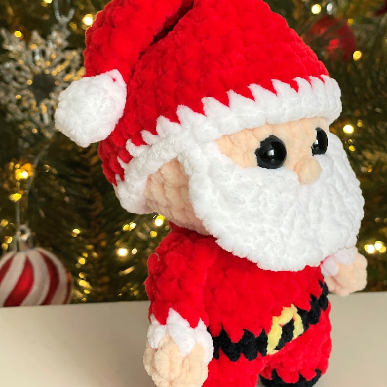 May include: A crocheted Santa Claus doll made of soft red, white, and black yarn. The figure features a red hat with a white pom-pom, a white beard, black safety eyes, and a black belt with a yellow buckle, all crafted in a plush texture.