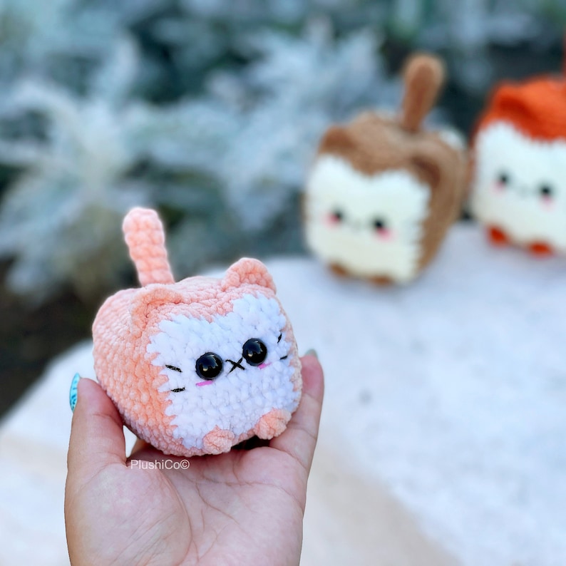 May include: A small, crocheted pink and white cat with black eyes. The cat is sitting in a hand.