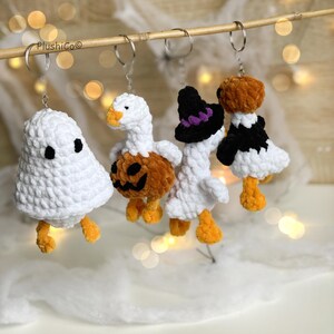 May include: Four handmade Halloween-themed keychains. The keychains include a white ghost, a goose holding a carved pumpkin, a goose wearing a witch hat, and a goose in a black and white outfit. Each keychain has orange feet and is made of a soft, textured material.