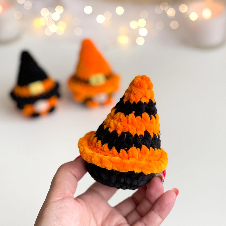 May include: A small, plush, crocheted witch hat featuring alternating horizontal stripes of bright orange and black yarn. The hat has a wide, rounded brim and a pointed top, crafted from a soft, fuzzy material. Two additional similar hats are visible in the background.