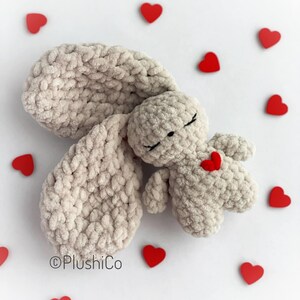 May include: A plush gray crocheted bunny with closed eyes and a red heart on its chest. The bunny is surrounded by red hearts on a white background.