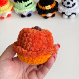 May include: A small, round, orange crocheted pumpkin figure with a textured surface and a brown stem on top. In the background, four additional crocheted figures in various colors and designs are visible, including green, black, and white patterns.