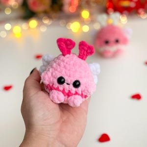 Peut inclure: Une petite peluche ronde crochet&eacute;e en fil rose doux. Elle pr&eacute;sente deux yeux en plastique noirs et ronds, une minuscule bouche brod&eacute;e, deux antennes rose vif sur le dessus et de petites ailes blanches sur les c&ocirc;t&eacute;s. Une seconde peluche similaire est visible en arri&egrave;re-plan.