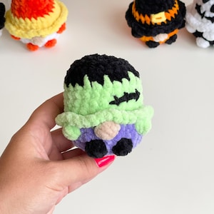 May include: A small, plush, crocheted figure resembling a monster with a black top, light green face, stitched scar detail, and a purple base. The figure has a small beige nose and two black feet. Other similar crocheted figures are visible in the background.
