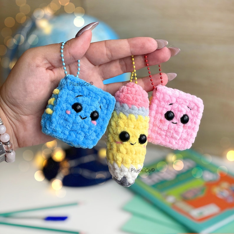 May include: Three handmade crochet keychains: a blue square book, a yellow pencil with a pink eraser, and a pink square. Each keychain has a smiling face and a metal chain for attachment. The keychains are from Plushico&copy;.