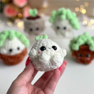 May include: A small, round, white crocheted plush toy with a soft, textured surface, black plastic eyes, and pink embroidered cheeks. It features a tiny green sprout on top. Other similar crocheted plant-themed plush toys are visible in the background.