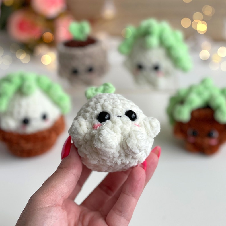 May include: A small, round, white crocheted plush toy with a soft, textured surface, black plastic eyes, and pink embroidered cheeks. It features a tiny green sprout on top. Other similar crocheted plant-themed plush toys are visible in the background.