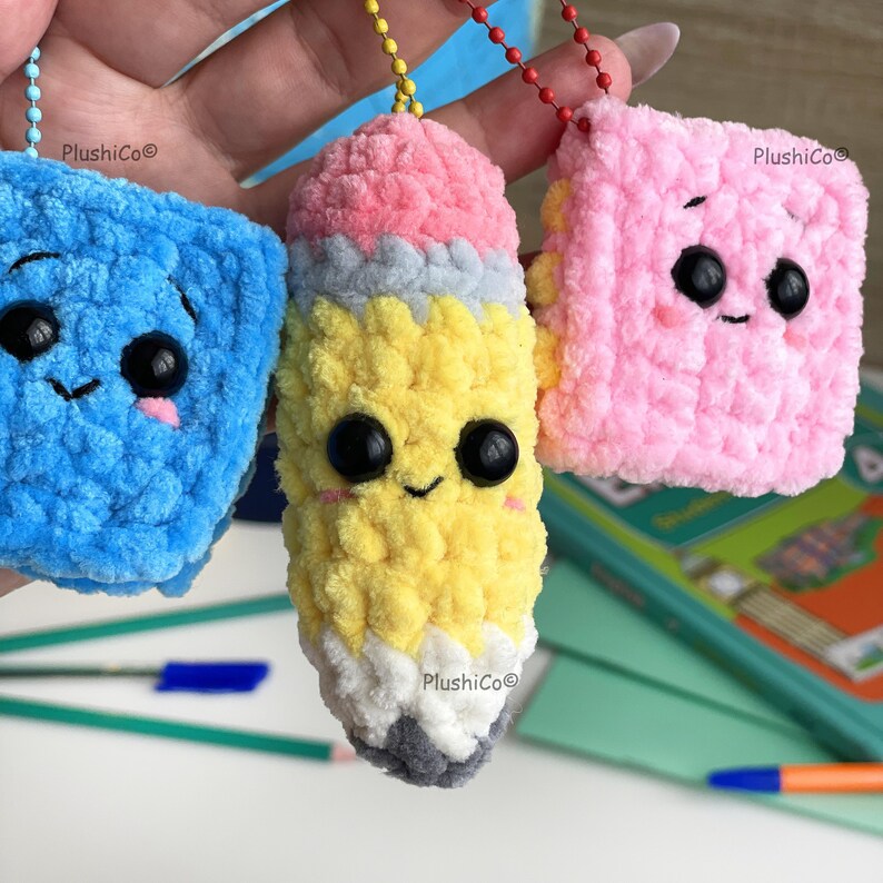 May include: Three plush keychains: a blue square, a yellow pencil, and a pink square, each with a smiling face and black eyes. The pencil has a pink eraser, gray tip, and the text "PlushiCo".