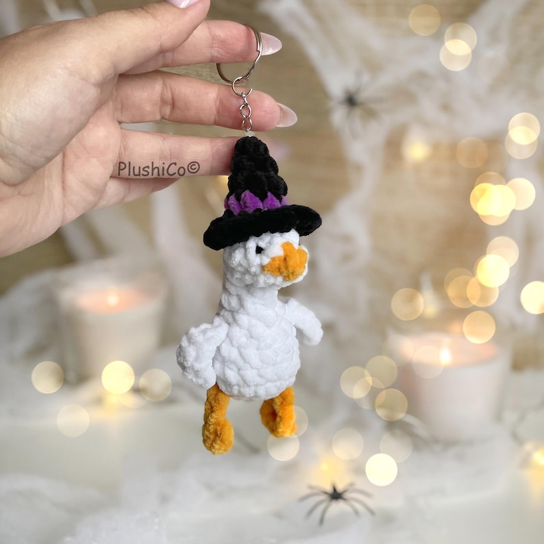 May include: A white crochet goose keychain wearing a black and purple witch hat. The goose has orange feet and beak. The keychain is held by a hand. The background has bokeh lights and spider decorations.