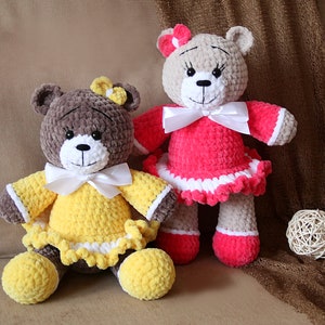 May include: Two crocheted teddy bears wearing dresses. The bear on the left is wearing a yellow dress with a white bow and the bear on the right is wearing a pink dress with a white bow.