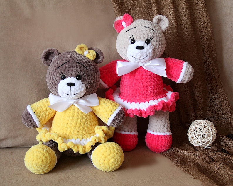 May include: Two crocheted teddy bears wearing dresses. The bear on the left is wearing a yellow dress with a white bow and the bear on the right is wearing a pink dress with a white bow.