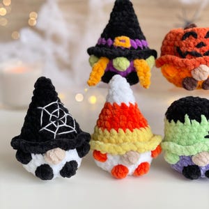 May include: A collection of five small, plush, crocheted figures featuring Halloween themes. The figures include a black witch hat gnome, a pumpkin, a gnome with a spiderweb hat, a candy corn gnome, and a green monster with black hair.
