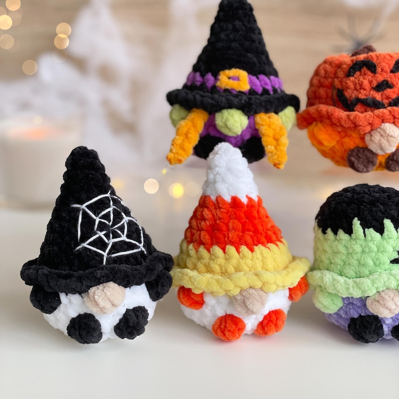 May include: A collection of five small, plush, crocheted figures featuring Halloween themes. The figures include a black witch hat gnome, a pumpkin, a gnome with a spiderweb hat, a candy corn gnome, and a green monster with black hair.
