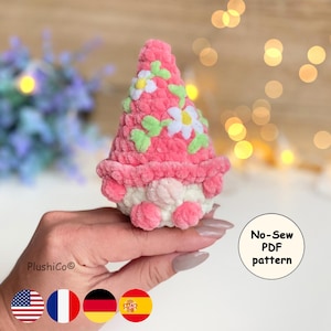 Puede incluir: Un gnomo de ganchillo rosa y blanco hecho a mano con un gorro cónico decorado con flores blancas y hojas verdes. El gnomo tiene una cara redonda blanca y una barba rosa. El texto "No-Sew PDF pattern" es visible.