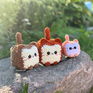 May include: Three crocheted cat plushies in different colours and sizes. The largest plush is orange and white, the middle plush is brown and white, and the smallest plush is pink and white.
