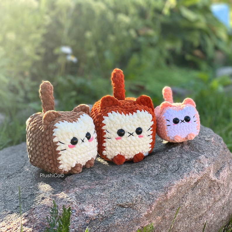 May include: Three crocheted cat plushies in different colours and sizes. The largest plush is orange and white, the middle plush is brown and white, and the smallest plush is pink and white.