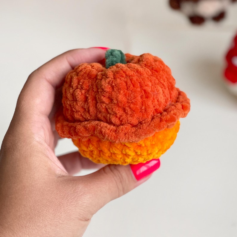 May include: A small, round, crocheted pumpkin made from soft, textured orange yarn. The pumpkin features a distinct, ruffled upper section in a darker shade of orange and a small, dark green stem at the very top, creating a layered, plush appearance.