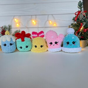 May include: A set of five small crocheted plush ornaments featuring a light blue candle, a mint green gift box with a red bow, a yellow bell with a pink bow, a pink mitten, and a light blue winter hat with a white pom-pom, all with embroidered faces.