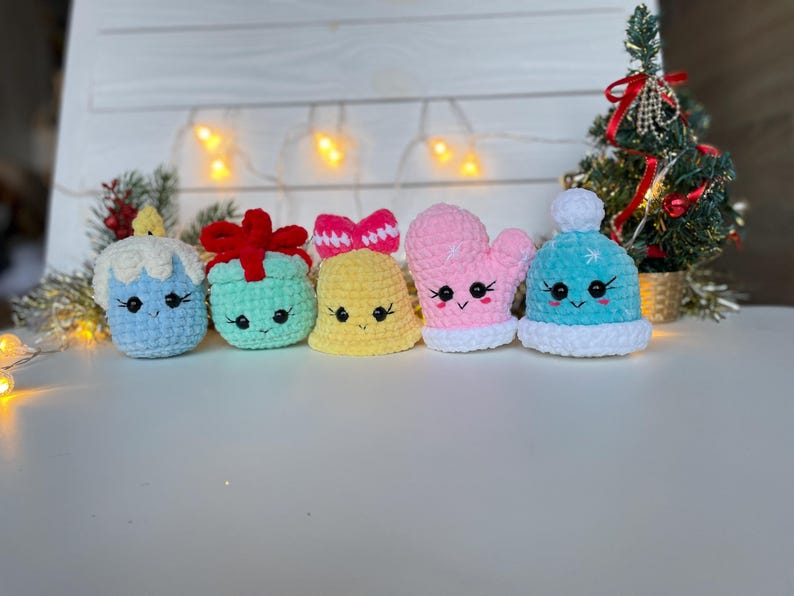May include: A set of five small crocheted plush ornaments featuring a light blue candle, a mint green gift box with a red bow, a yellow bell with a pink bow, a pink mitten, and a light blue winter hat with a white pom-pom, all with embroidered faces.