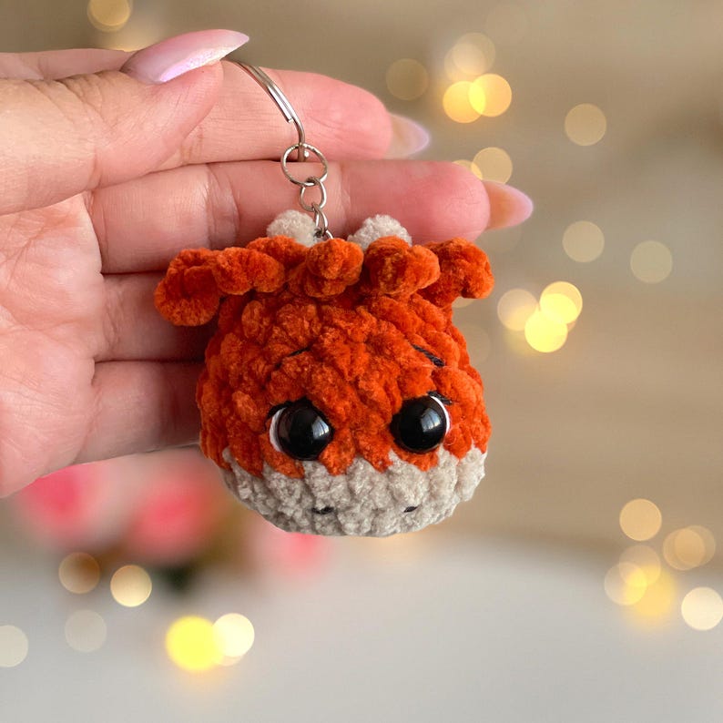May include: A handmade orange and tan crocheted fox keychain with large black eyes. The fox has a fluffy texture and a silver keyring attached to its head. The background is blurred with bokeh lights.
