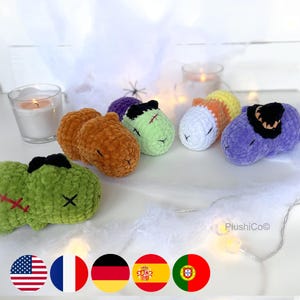 Puede incluir: Cinco peluches de crochet con diferentes temas de Halloween. Los peluches tienen forma de calabaza, fantasma, zombie, bruja y vampiro. Todos los peluches están tumbados sobre una superficie blanca.