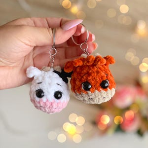 May include: Two handmade crochet keychains. One is a white cow with black spots and a pink muzzle, the other is an orange fox. Both have large black eyes and are attached to silver keyrings.